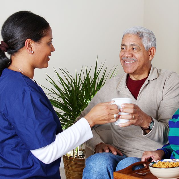 Profissional de saúde entrega uma xícara para um idoso sorridente em ambiente domiciliar, representando cuidado humanizado e conforto no home care.