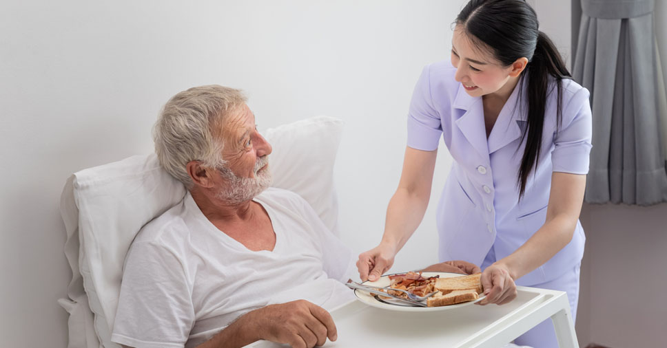 Cuidadora auxiliando um idoso acamado durante a refeição, em ambiente domiciliar, representando cuidado integral, rotina e atenção à saúde física e emocional.